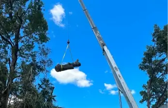 A large commercial crane lifting a heavy section of a cut tree log high in the air using yellow rigging straps against a bright blue sky with sparse white clouds.