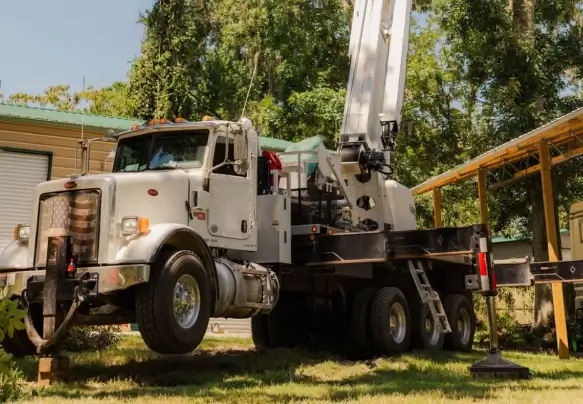 A large white commercial crane truck lifted completely off its front wheels by heavy-duty outrigger stabilizer jacks on a grassy lawn next to buildings and trees.